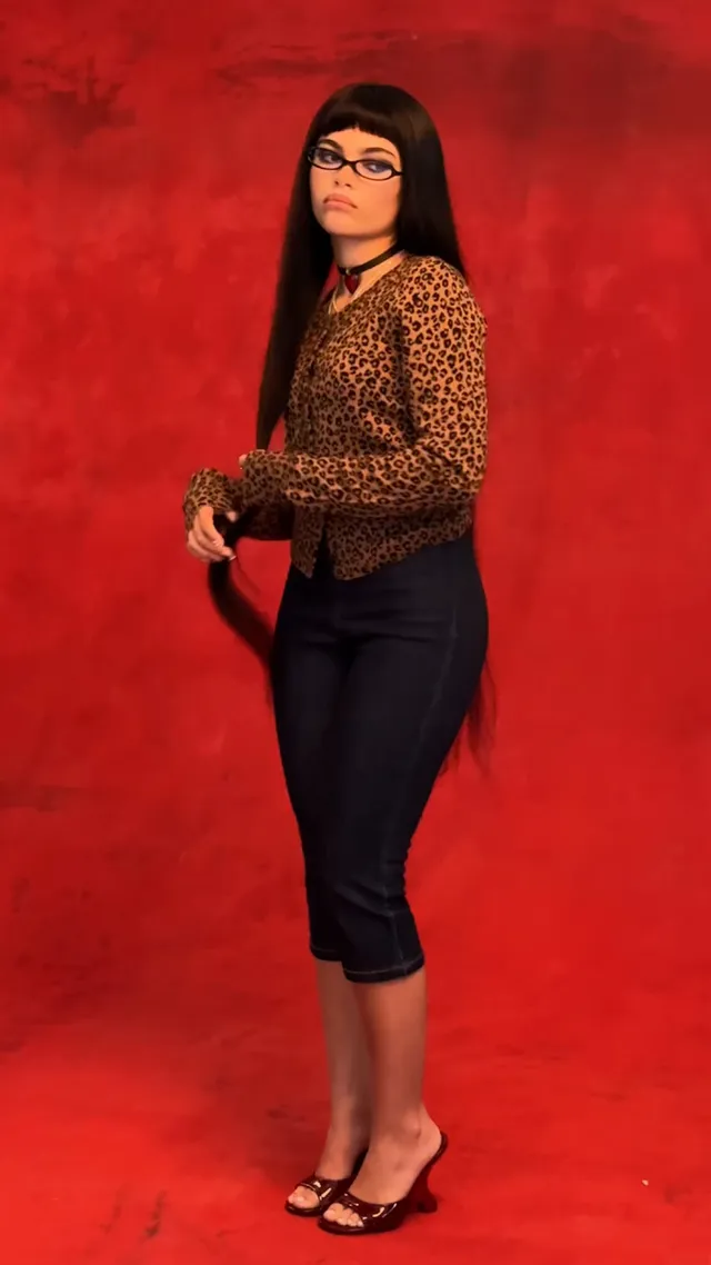 Ariana Greenblatt standing in a patterned top and flat sandals against a red backdrop.