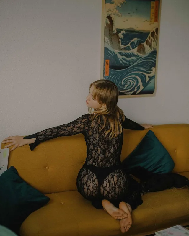 Clara Galle kneeling barefoot on a yellow sofa in a black lace dress.