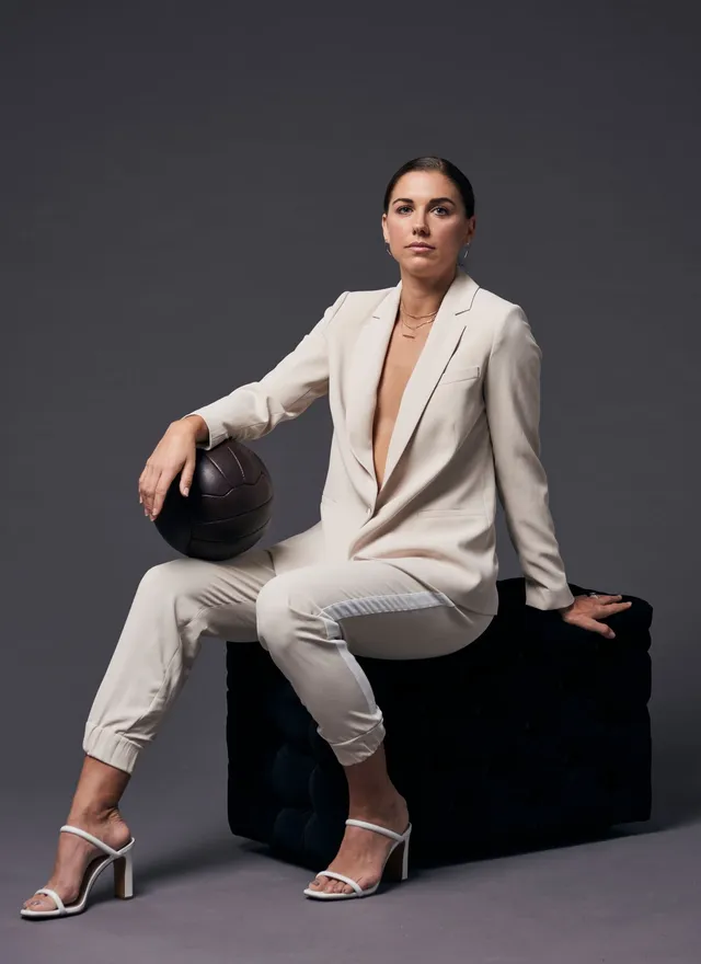 Alex Morgan sitting in silver heeled sandals and a pale suit during a studio portrait.