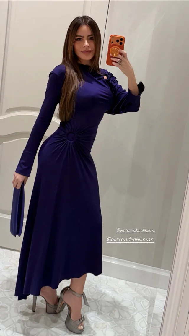 Sofía Vergara posing for a mirror photograph in a purple dress and platform heels.