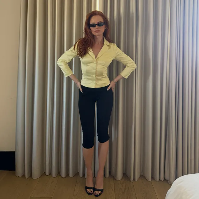 Madelaine Petsch posing in a yellow blouse and black trousers with heels.