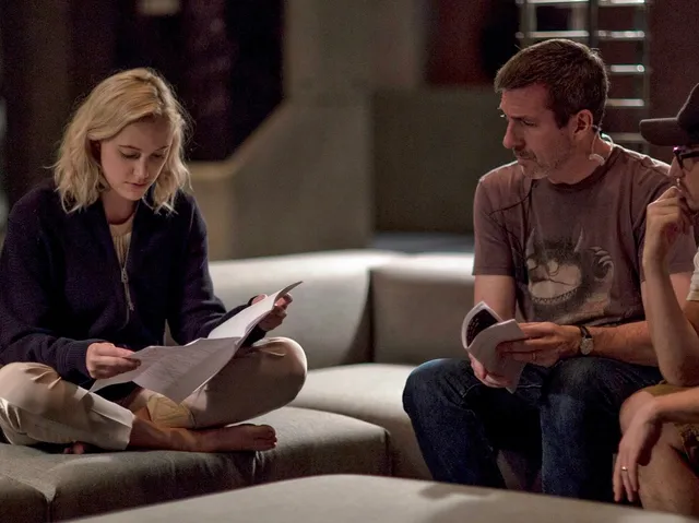 Maika Monroe sitting barefoot cross-legged on a sofa in a scene.