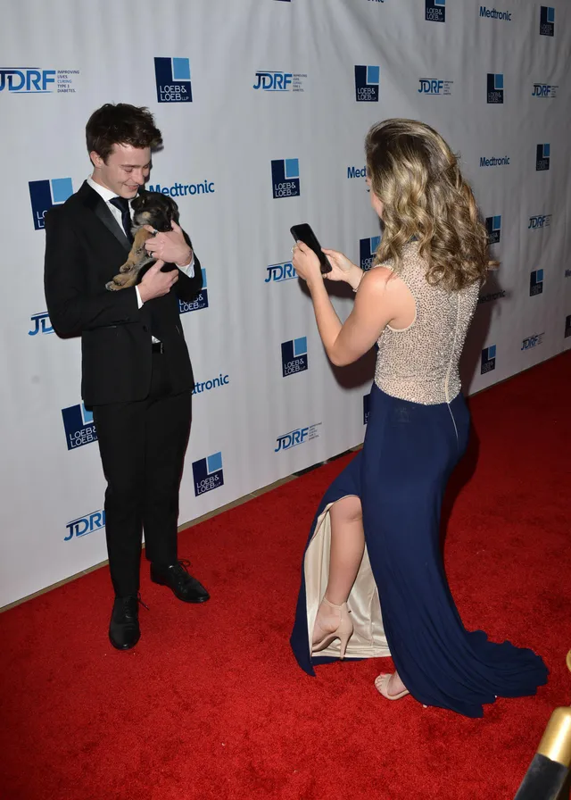 Brec Bassinger taking a photograph at an event in a long gown and heels.