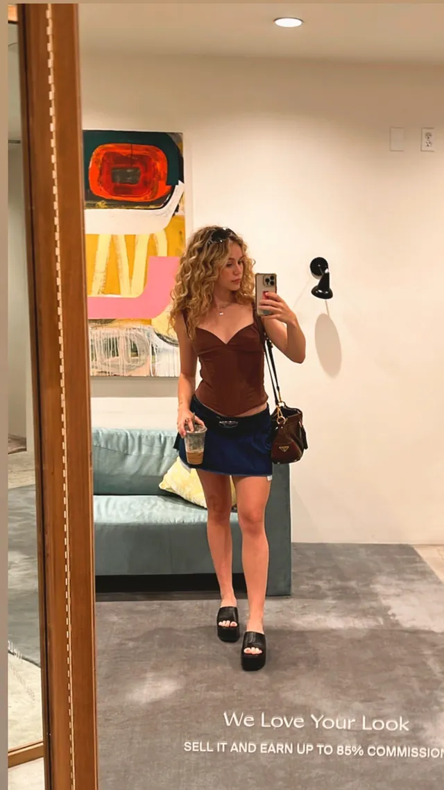 Brec Bassinger taking a mirror selfie in a brown top, denim shorts and dark loafers.
