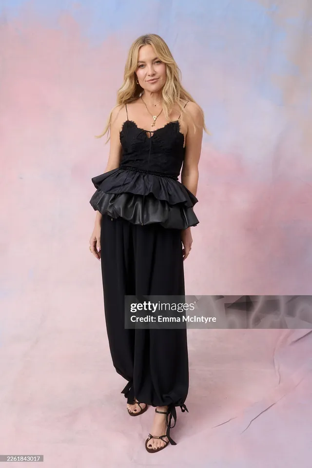 Kate Hudson posing against a pale studio backdrop in a black dress.
