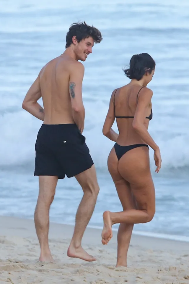 Bruna Marquezine walking barefoot beside a man on a beach in a black bikini.