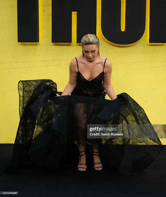 Florence Pugh sitting in a black dress and heels at an event.