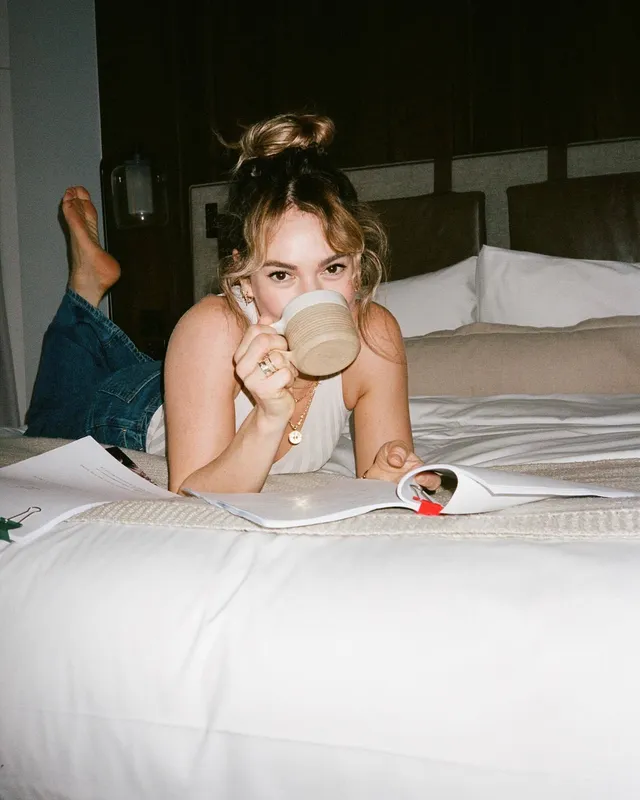 Lily James lying on a bed with a mug and one bare foot raised.