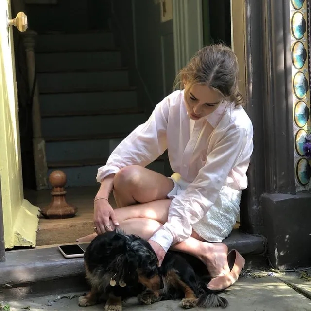 Emilia Clarke sitting barefoot on steps beside a black dog.