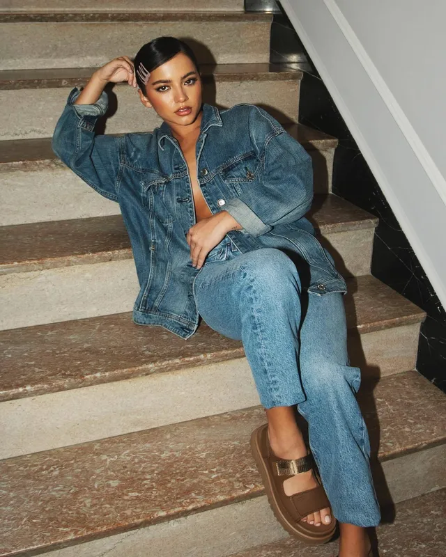 Isabela Merced sitting on stairs in denim and brown platform sandals.