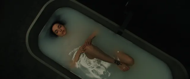 Dove Cameron reclining barefoot in a dark bathtub.