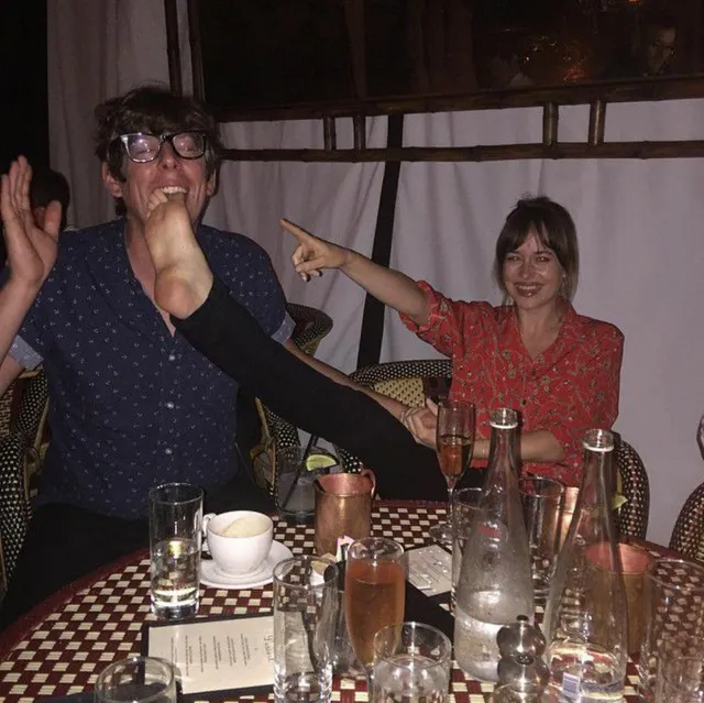 Dakota Johnson holding up a foot during a dinner-table photograph.