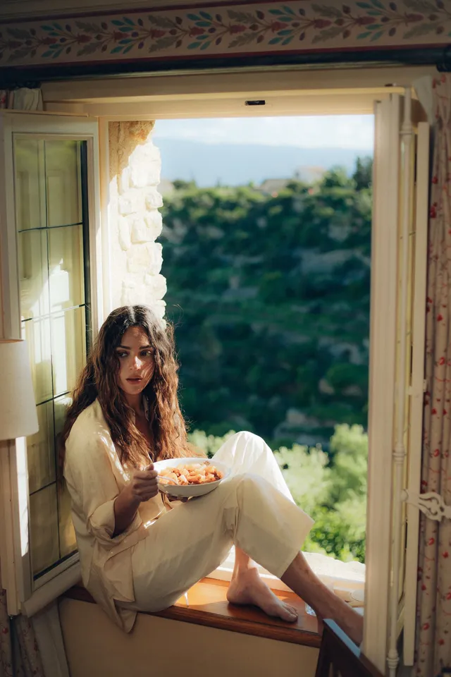 Adria Arjona sitting barefoot in an open window while holding a plate.