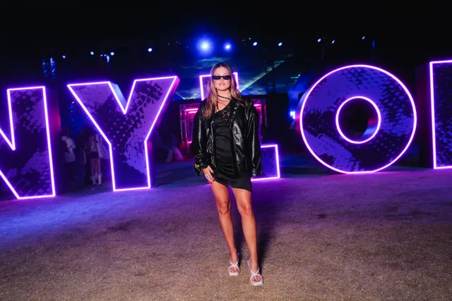 Olivia Holt posing in a black outfit and white flat sandals before an illuminated display.