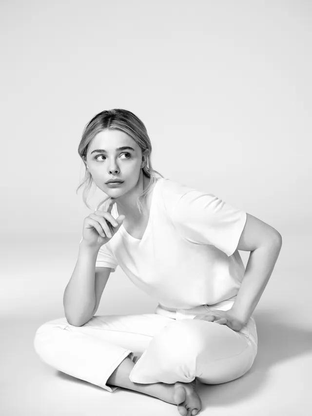 Chloë Grace Moretz sitting barefoot in a white outfit in a monochrome portrait.