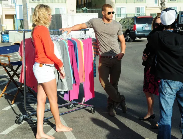 Kate Upton standing barefoot beside clothing racks outdoors.