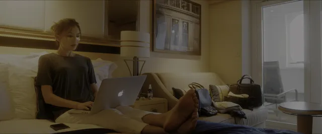 Gemma Chan using a laptop with her feet resting on a table in a screen scene.