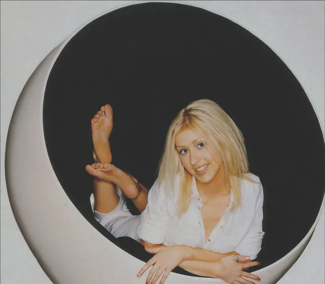 Christina Aguilera sitting in a circular window with a bare sole raised.