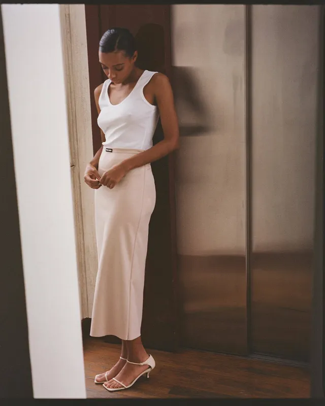 Sophie Wilde standing by a mirror in pale trousers and heeled sandals.