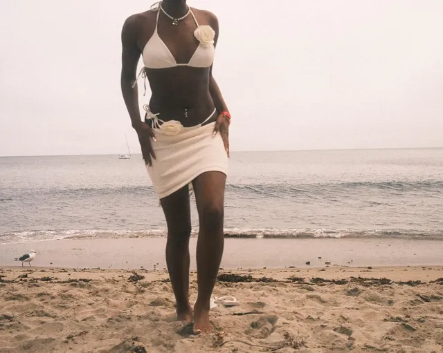Carlacia Grant standing barefoot on a beach in a white skirt and bikini top.