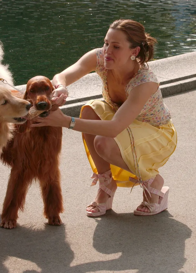 Jennifer Garner crouching beside a goat in a yellow skirt.