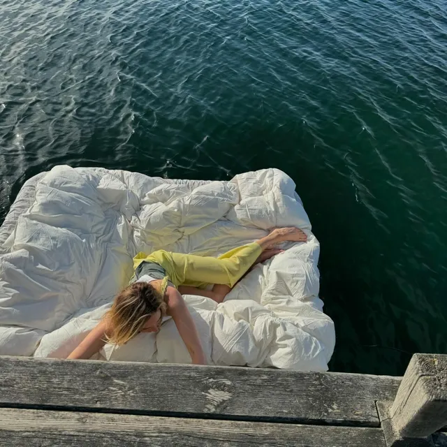 Rebecca Ferguson reclining in a yellow outfit on a mattress floating beside a stone ledge.