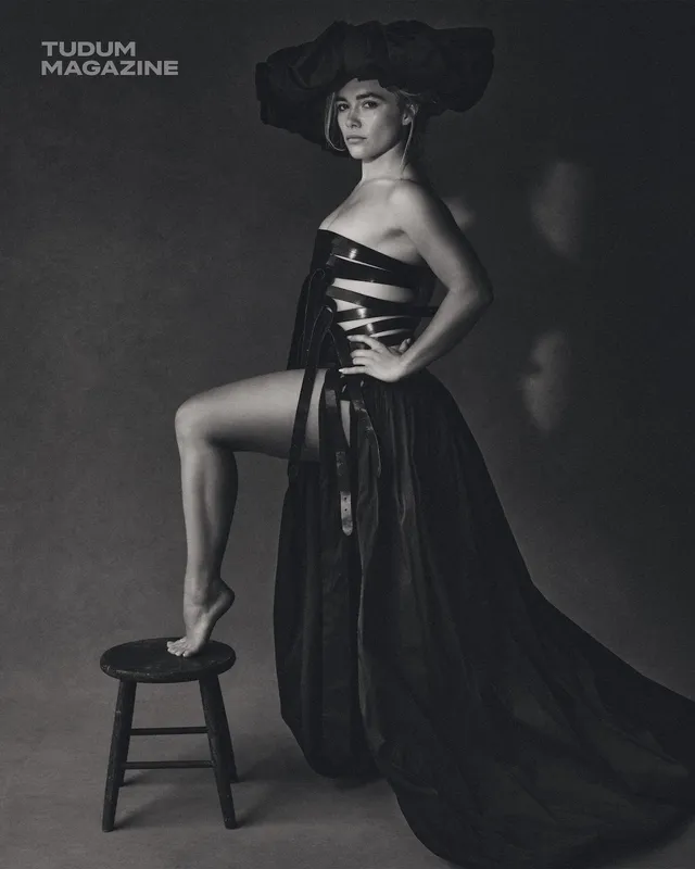Florence Pugh posing with one heel resting on a stool in a black outfit.