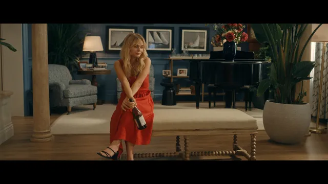 Emily Alyn Lind sitting on a stool in a red dress with bare feet visible in a scene.