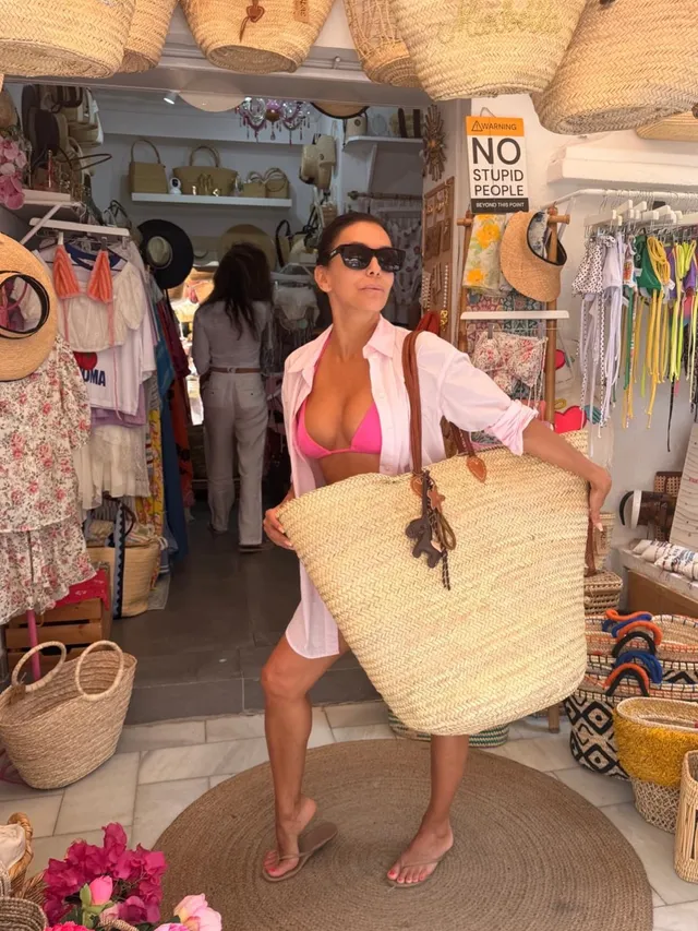 Eva Longoria standing inside a shop carrying a woven bag.
