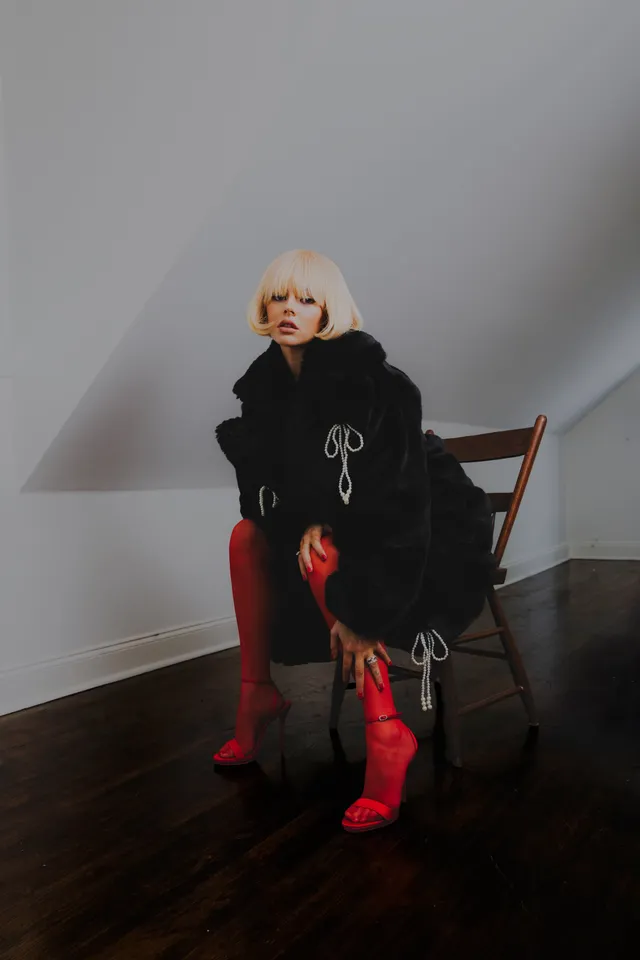Samara Weaving sitting in a dark coat and red heeled boots.