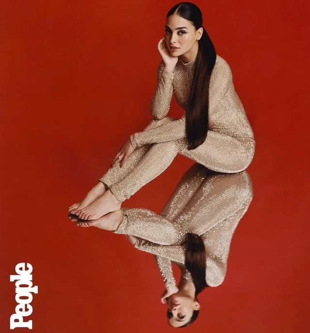 Melissa Barrera sitting on a reflective floor in a gold outfit.