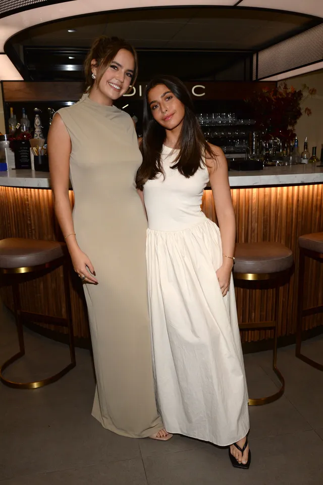 Bailee Madison posing with a friend in a long beige dress and clear heels.