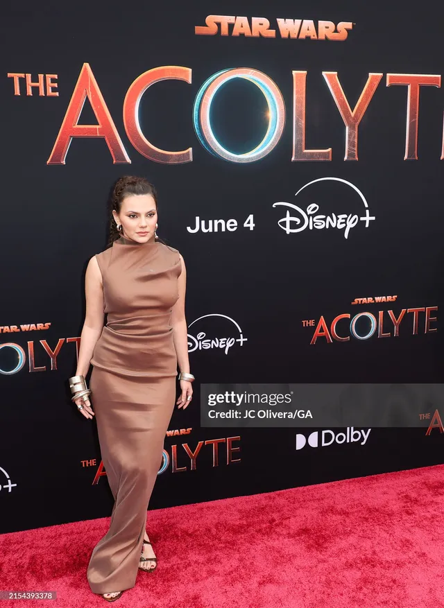 Dafne Keen standing on a premiere carpet in a long brown dress and heels.