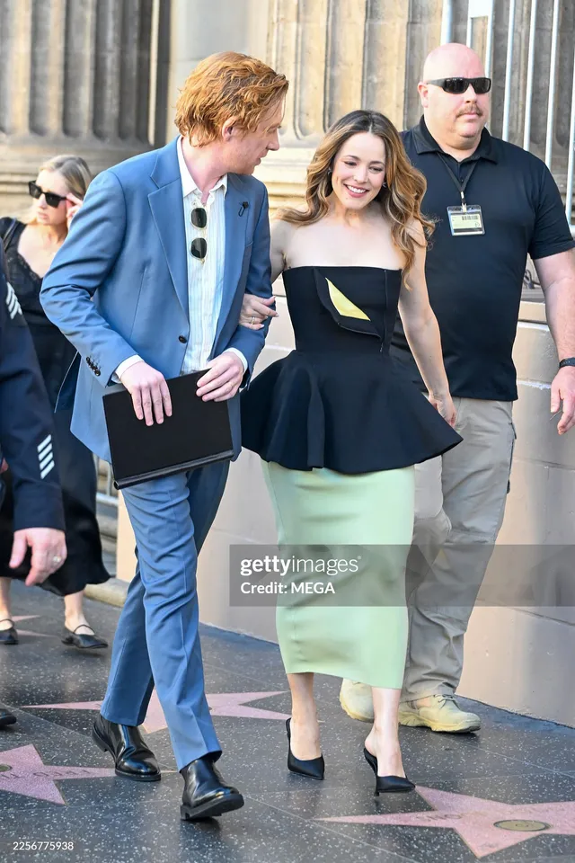 Rachel McAdams walking beside other people in a black top and pale skirt with heels.