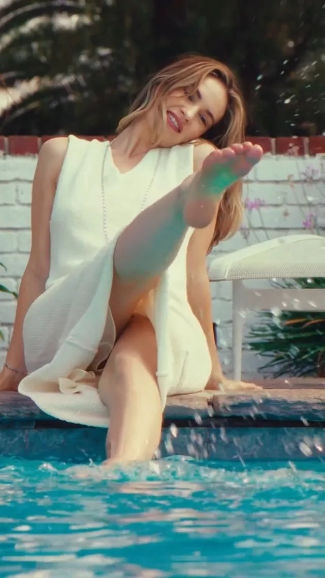 Lily James sitting by a pool with one bare sole raised.