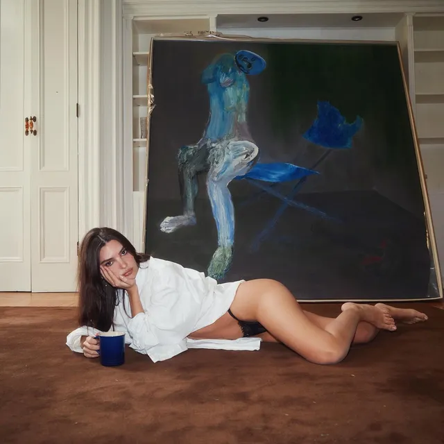 Emily Ratajkowski lying barefoot on a rug beside a painting.