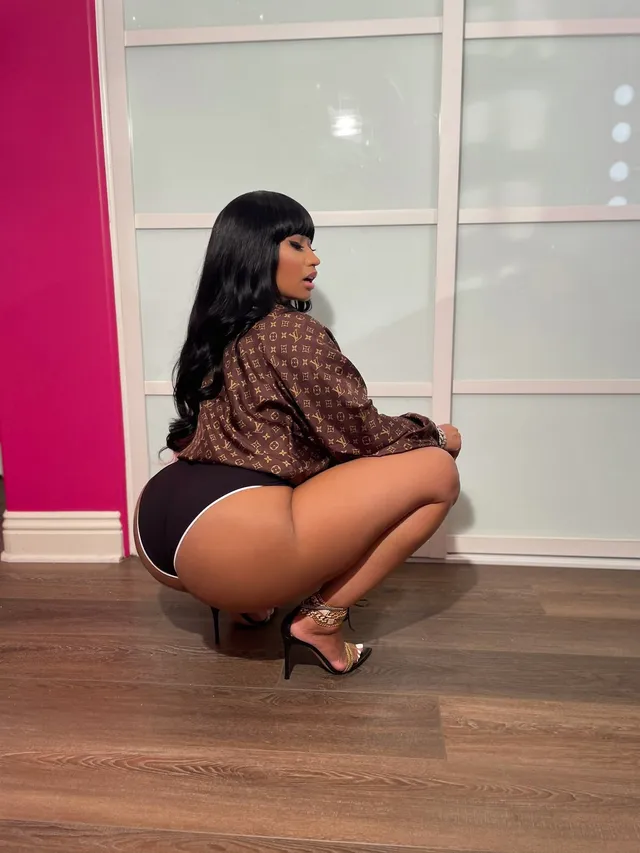 Nicki Minaj crouching indoors in a patterned top and black heels.
