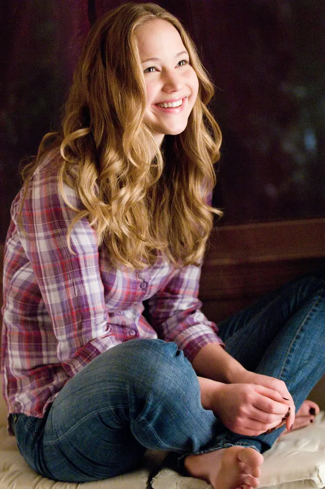 Jennifer Lawrence sitting indoors in a checked shirt and jeans.