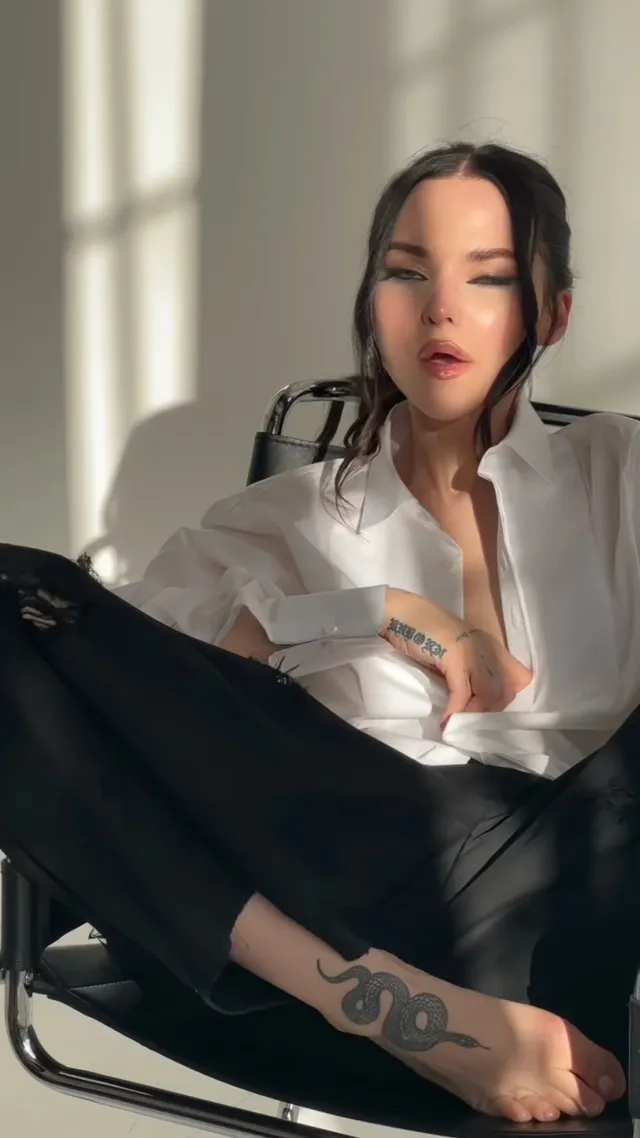 Dove Cameron sitting barefoot in a chair by a window.