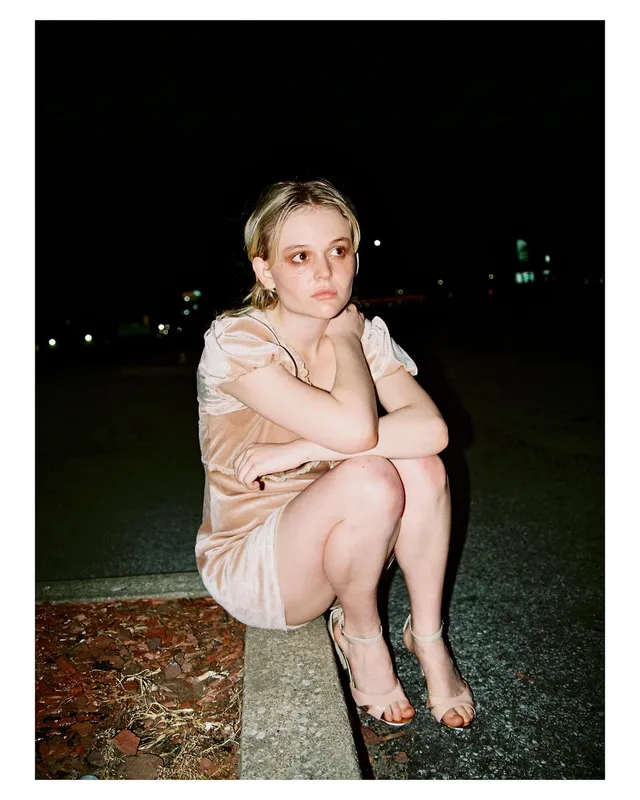 Emily Alyn Lind crouching outdoors in a pale dress and strappy heels at night.