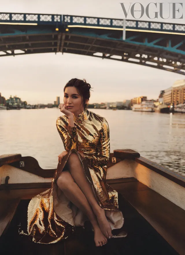 Gemma Chan sitting on a boat in a patterned gold outfit.