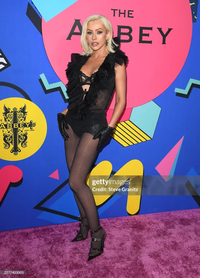 Christina Aguilera standing at an event in a black outfit and lace-up heels.
