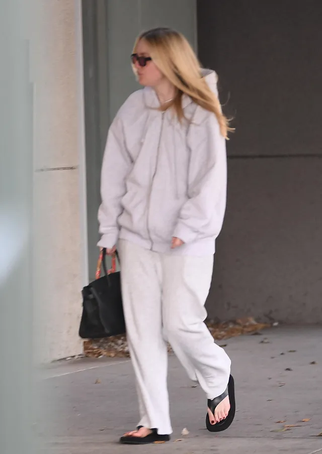 Dakota Fanning walking in a pale sweatshirt, trousers and black sandals.