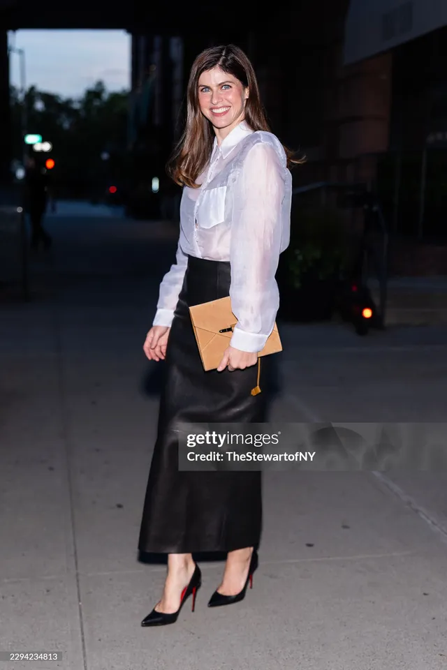 Alexandra Daddario posing outside in a white blouse, dark skirt and pointed black heels.