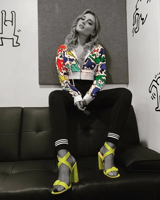 Meg Donnelly sitting on a sofa in dark trousers, striped socks and bright sandals.