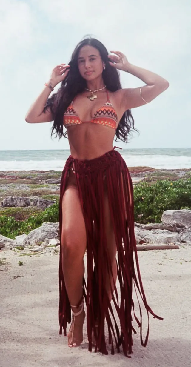 Emily Tosta standing barefoot by the sea in a fringed skirt.