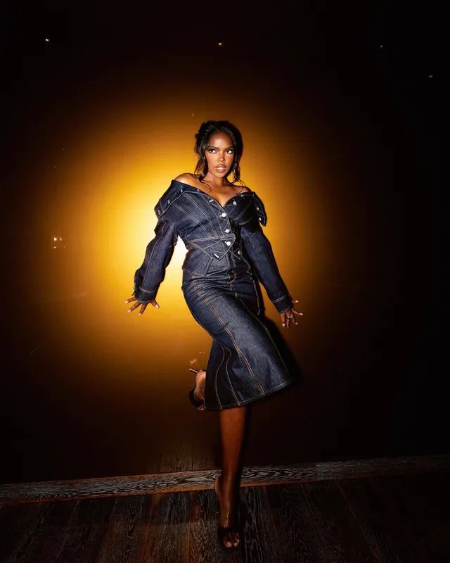Ryan Destiny posing in a denim dress and black heels against a warm backdrop.
