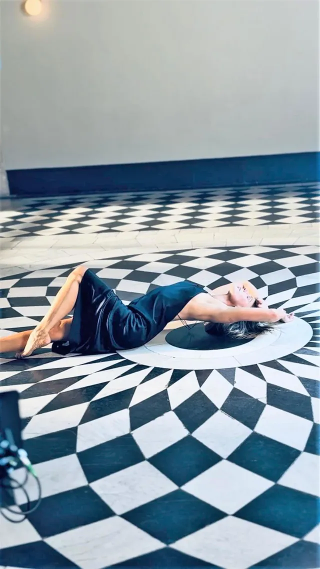 Rebecca Ferguson reclining barefoot in a dark dress on a patterned studio floor.
