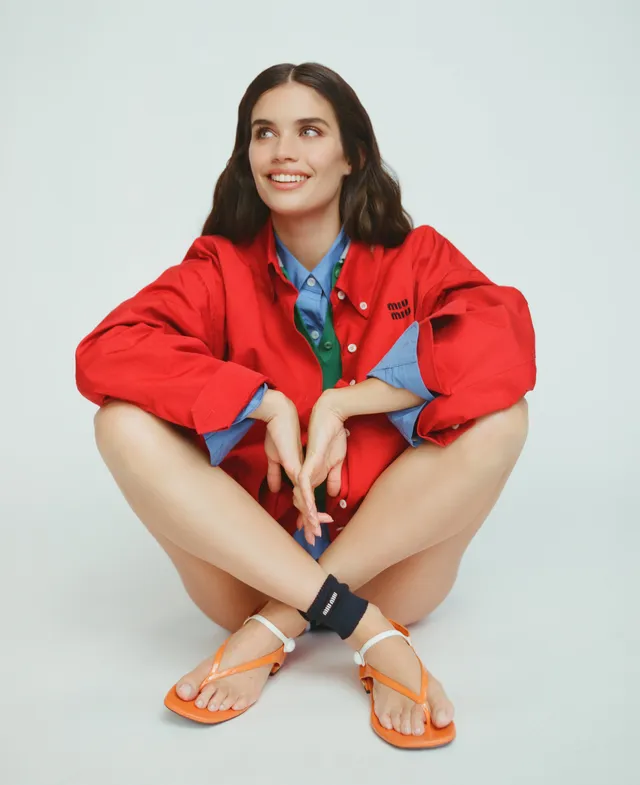 Sara Sampaio crouching in orange flat sandals and a red outfit during a studio shoot.