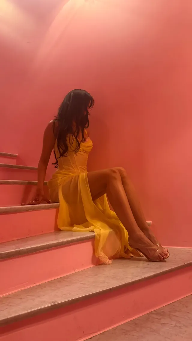 Camila Cabello sitting on pink stairs wearing a yellow dress and heels.